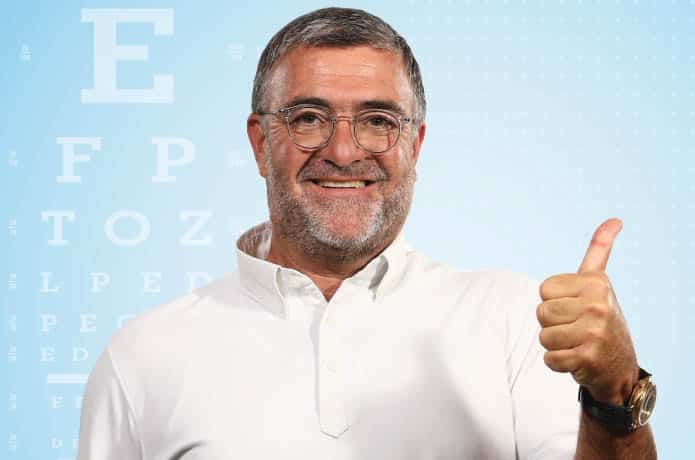 גבר מחייך עם משקפיים בחולצה לבנה מרים אגודל מול טבלת בדיקת ראייה, ומגביר את המודעות למיגרנה של העין.